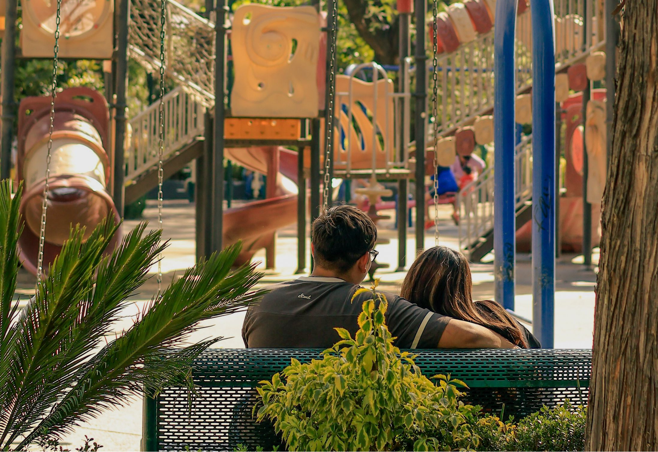 A man and woman sitting on a bench overlooking a playground in a local park
