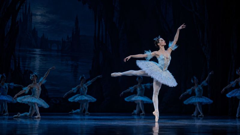 Ballet dancers performing on stage, featuring a ballerina in a light blue tutu dancing en pointe under soft blue lighting.