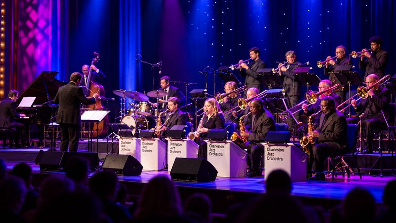 Charleston Jazz Orchestra performing live on stage, featuring saxophones, trumpets, trombones, and a conductor under blue and purple lighting.