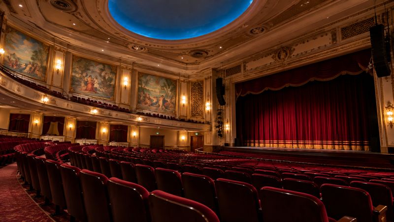 Elegant historic theater auditorium featuring red velvet seats, ornate balconies, decorative wall murals, a blue domed ceiling, and a stage with red curtains.