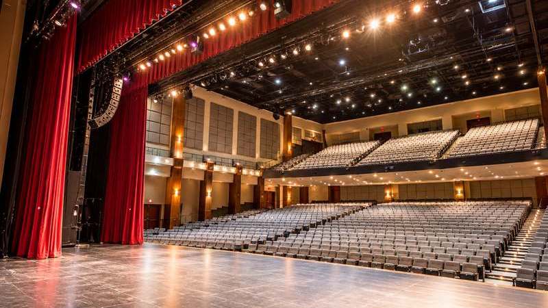 Empty performing arts theater viewed from the stage, with red curtains, rows of seats, and overhead stage lighting.