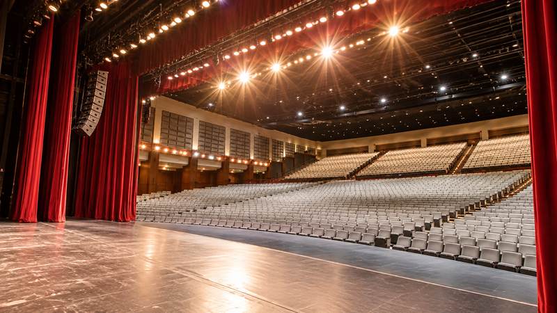 Empty theater auditorium viewed from the side of the stage, featuring red curtains, rows of seats, and bright stage lighting.