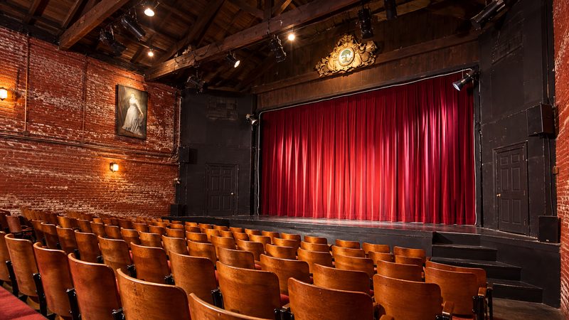Interior of Queen Street Playhouse in Charleston, featuring wooden theater seats, red stage curtains, exposed brick walls, and overhead lighting.