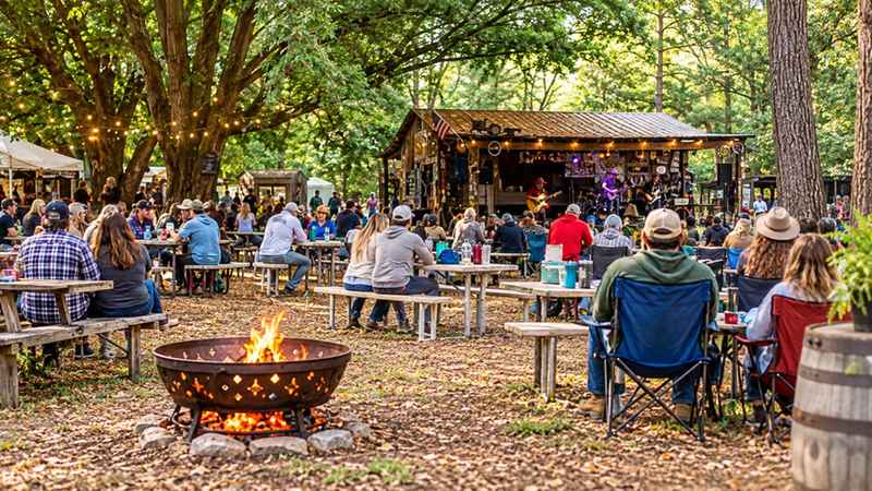 Outdoor live music gathering at Awendaw Green with a rustic stage, seated audience, trees, and a fire pit.